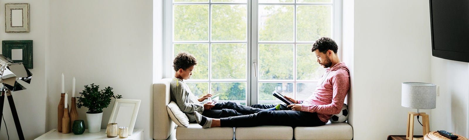 A father and son are relaxing by a large window at home, reading and playing games together.