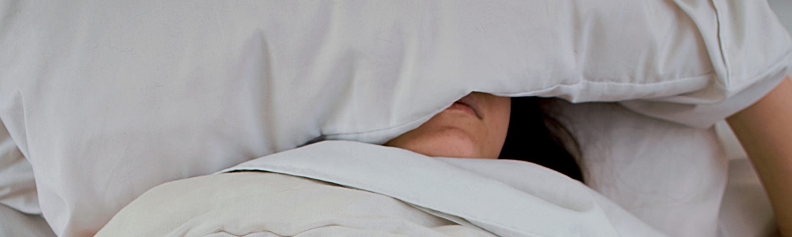 A girl is in bed, covering her head with a pillow. The blankets are pulled up to her chin, and she is holding her glasses in her other hand