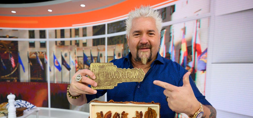 Guy Fieri holding and pointing to a small plaque that says “Flavortown”.