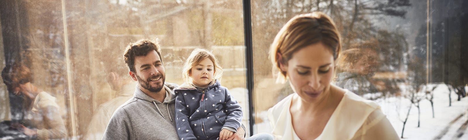 A photo of a family of four (mom, dad, two kids) sitting at home. Mom is on her computer and attending to a child.