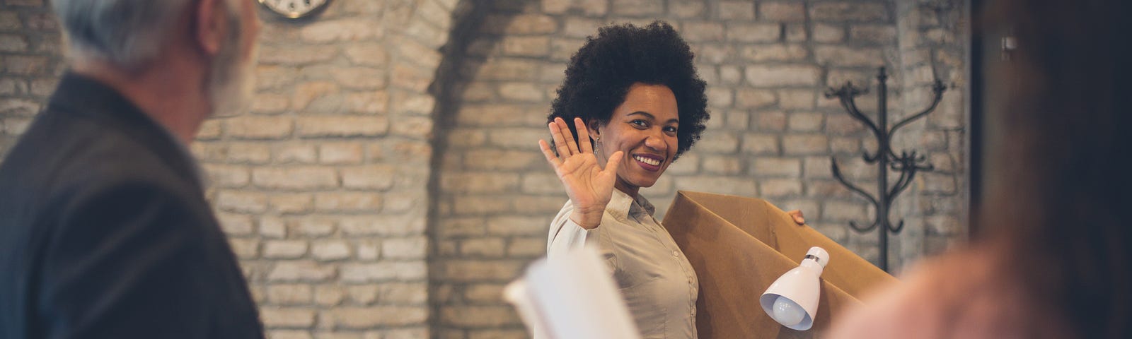 A woman smiles and waves goodbye to her coworkers, as she walks out with a box of her belongings.