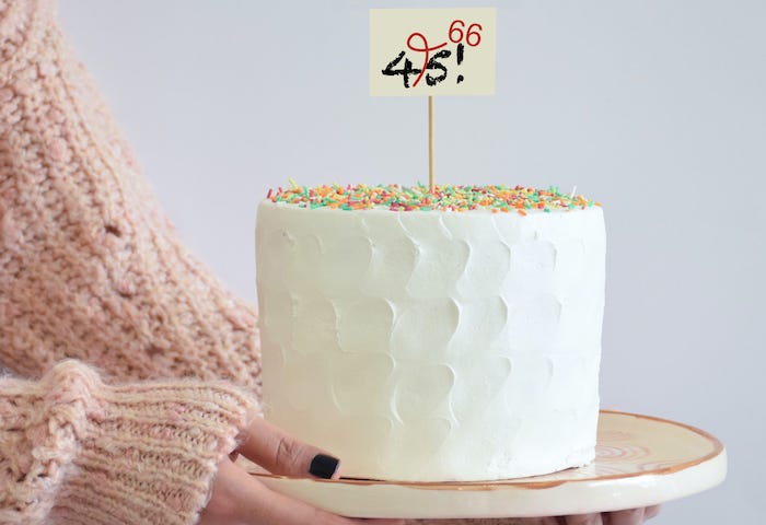 A woman’s arms holding a white birthday cake with a sign that says 45!, crossed out and corrected to 66.