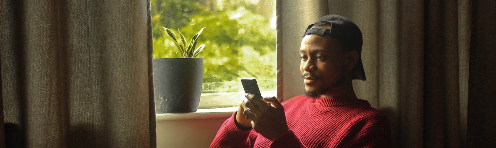 Black man rests by his window while looking at his phone.