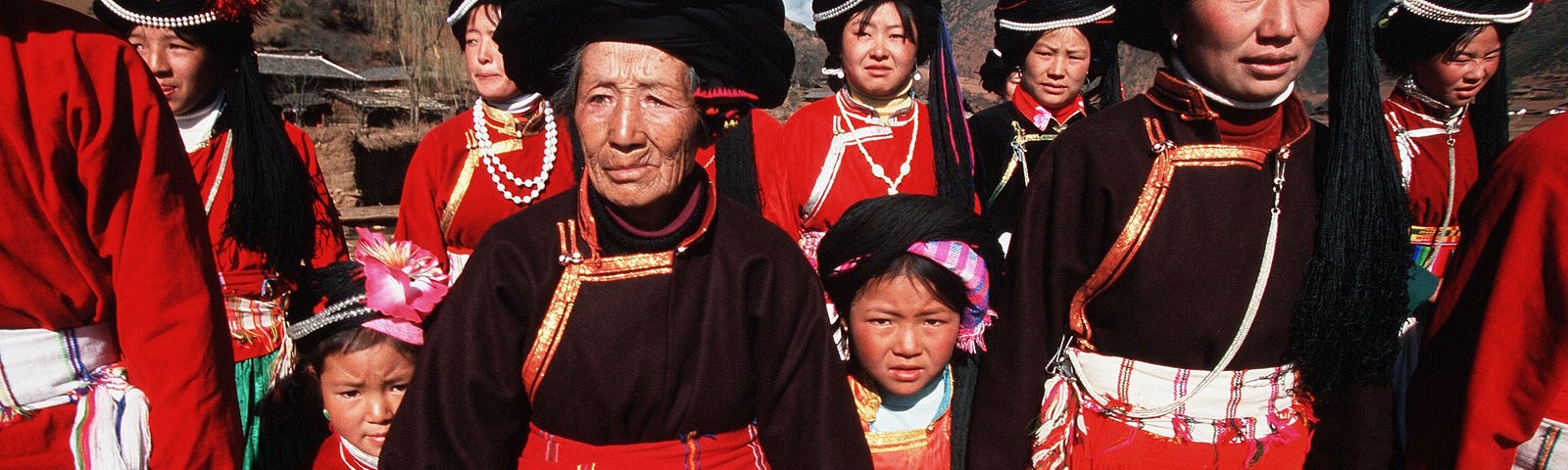 Mosuo women and girls, ranging from young kids to elderly women, wearing dresses with red, black, green, and white designs, holding hands outdoors.