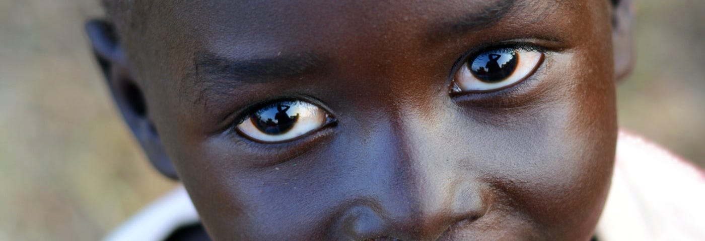 A close up of the face of a young African child in a loose white top, smiling directly at the camera.