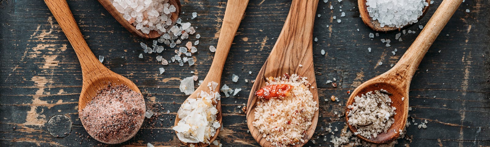 An assortment of different-sized wooden spoons on a weathered wooden surface; each heaped with a different color/texture salt