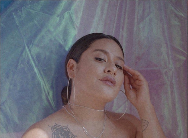 Amy Quichiz posing with her fingers held to her left temple in front of a backdrop of iridescent fabric.