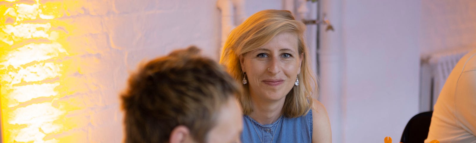 A photograph of a woman with blonde hair looking directly at the camera, smiling. In the foreground a table with colorful plastic pieces stacked together and a woman with short hair, not in focus.
