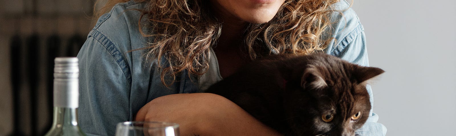 A woman with curly hair and glasses leans over a computer, smiling while holding a cat, with a bottle and glass of wine