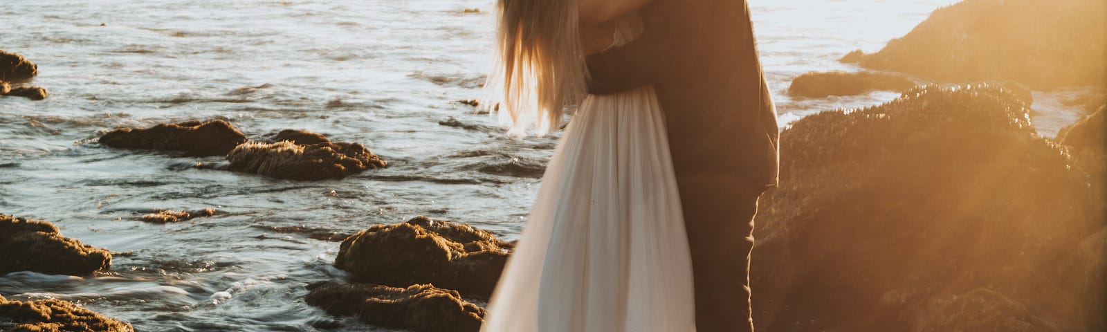 A tall man romantically clasps in his arms a long-haired woman in a flowing white dress. They’re kissing while standing on a rock on the beach, surrounded by water and mistiness to the horizon.