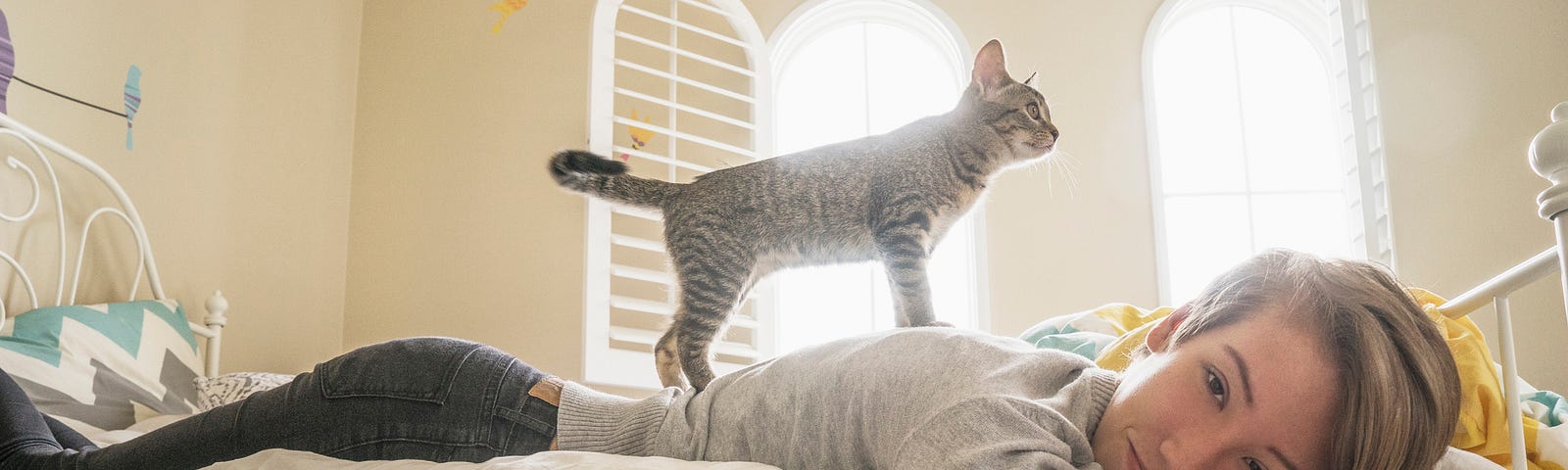 A young woman lies languidly in bed on her stomach while a cute cat stands on her back.