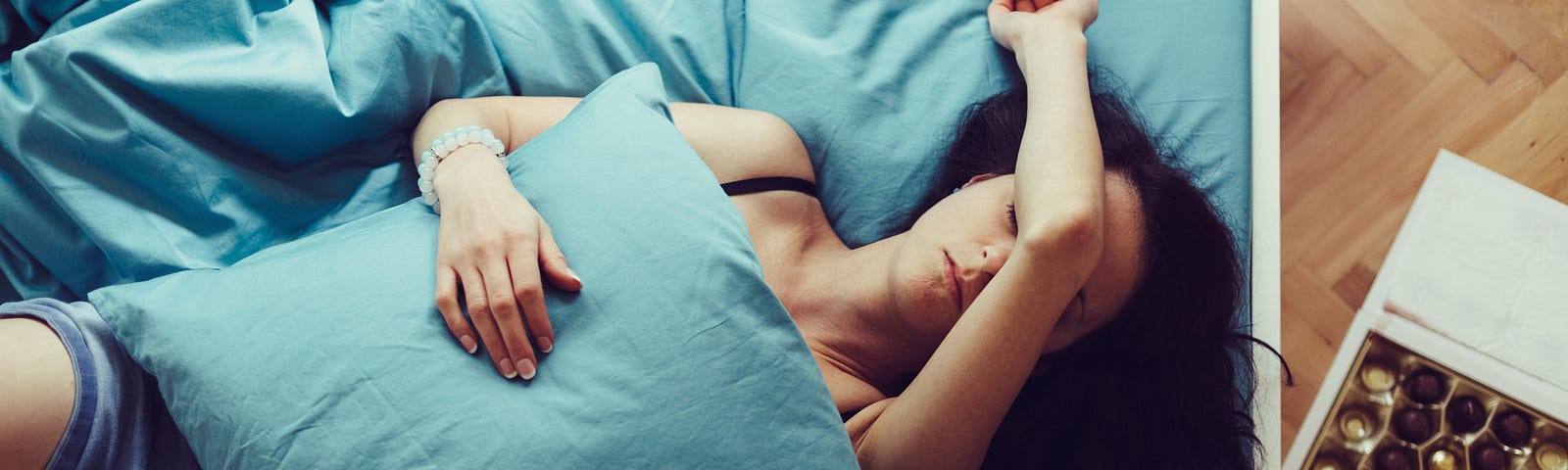 A tired woman lies on her bed with her arm covering her face and a pillow over her body.