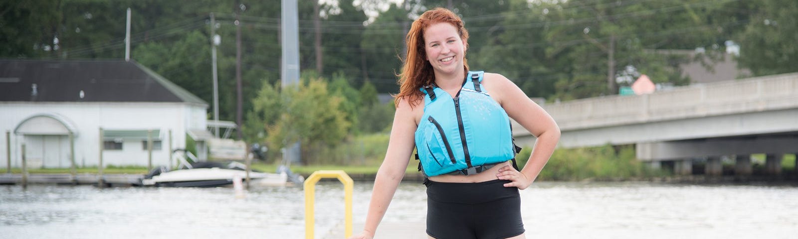 Woman wearing blue life jacket stands in front of a stand up paddle board on a dock