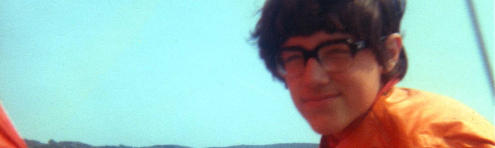 A young teenage boy in glasses, a red t-shirt, white pants and an orange life-jacket holds the tiller and the mainsail sheet of his wooden dinghy at sea, an area of open water and a headland behind him.