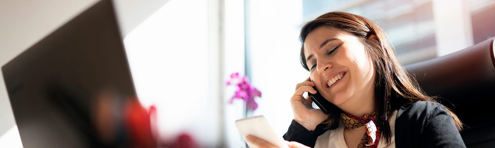 A photo of a smiling woman on the phone at her desk in front of her computer.