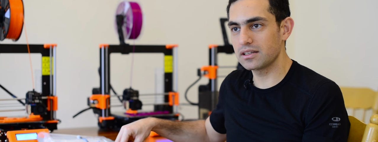 A man in a black shirt sitting next to three 3D printers on a desk.