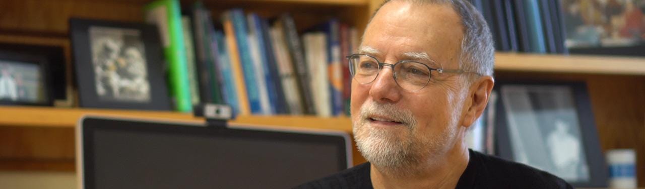 A middle-aged white man wearing glasses and a black turtleneck speaking in front of a bookcase.