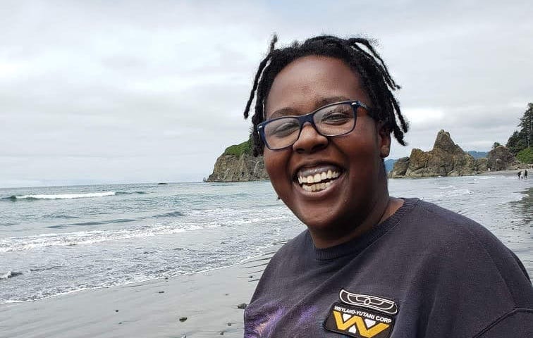 Short story and poetry collection by Aigner Loren Wilson article cover. Picture of author smiling on a beach. Behind her are rocky and tree ridden cliffs and the rolling ocean. Speculative fiction by Aigner Loren Wilson. Horror by Aigner Loren Wilson