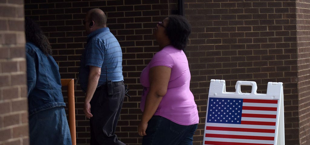 A photo of three early voters in Florida lining up at a poll. There is a “EARLY VOTING VOTACION ADELANTADA” sign.