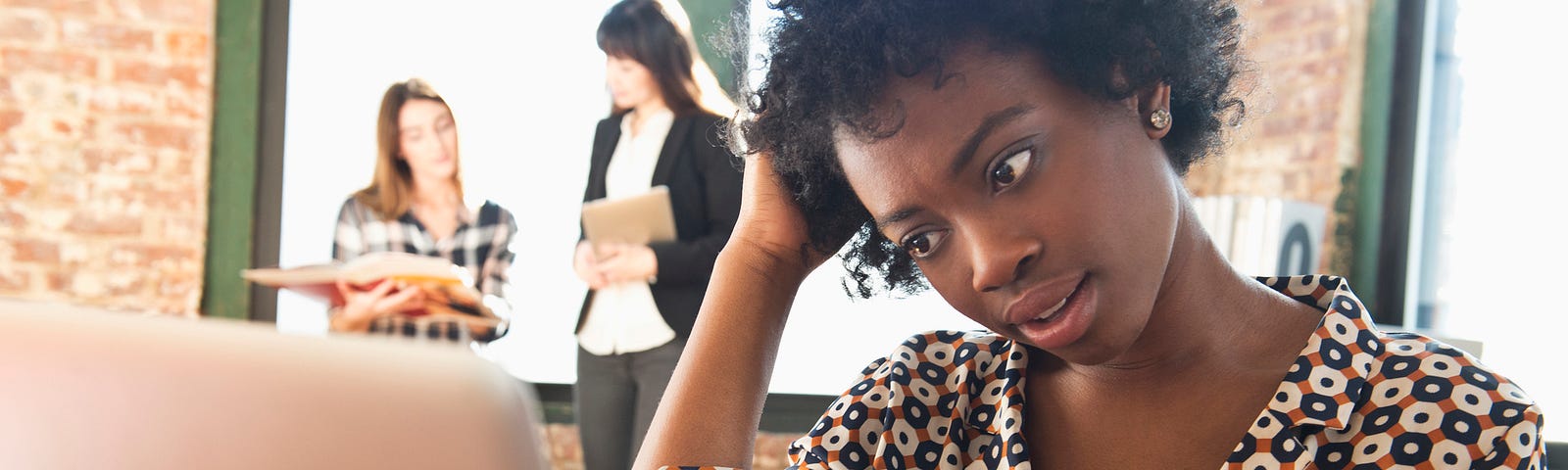 Black woman at work holding a phone while looking at her laptop.