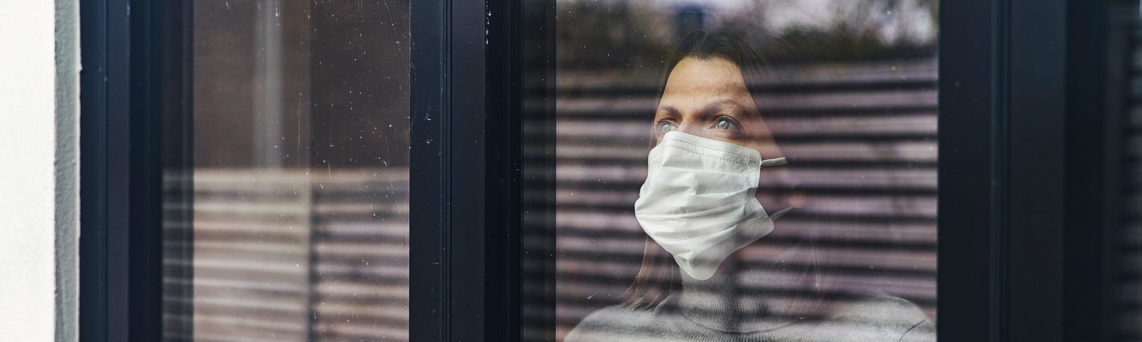 A photo of a woman wearing a mask looking out the window from her home.