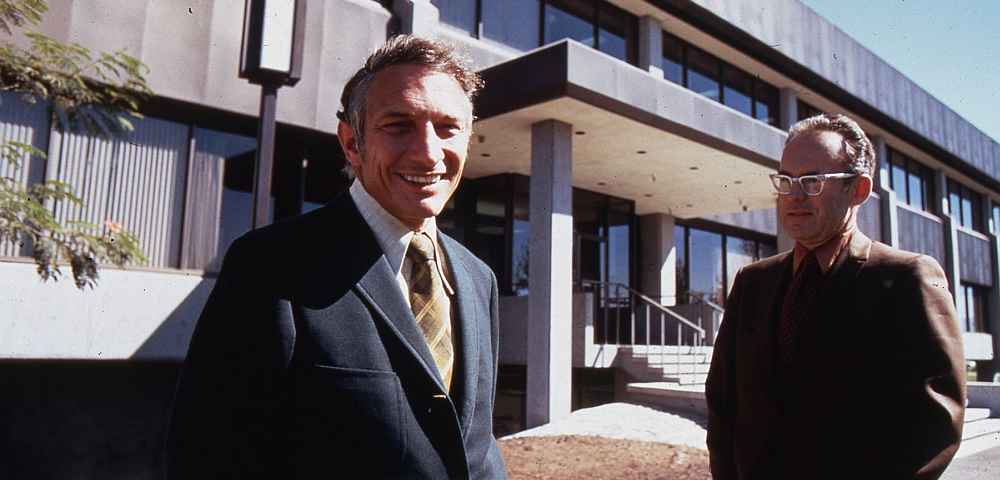 A vintage photo of two men in suits standing in front of a building on a college campus.