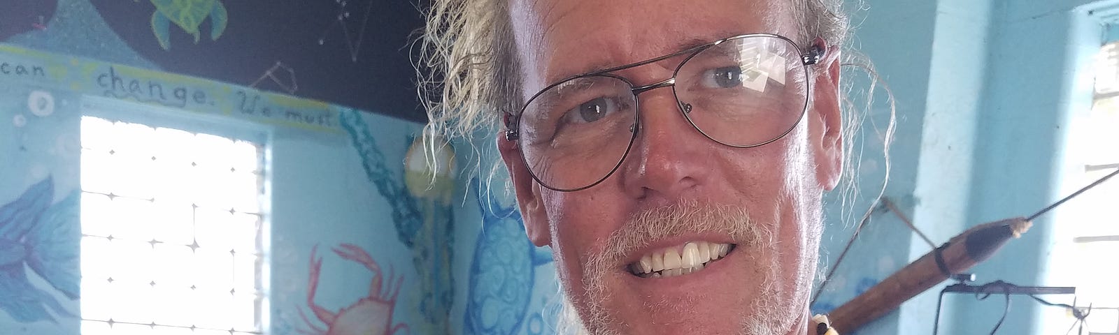 A smiling man in glasses, blond hair tied back and wearing a yellow T-shirt holds a Ball python snake, nestled in his cupped hands. In the background, an aquarium can be seen.