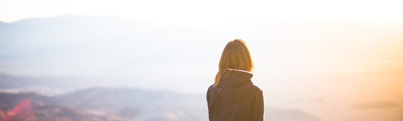 A woman in a black jacket and jeans sits at the top of a mountain, looking into the sunset. You can see the back of her head.