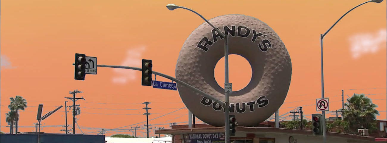 A photograph of Randy’s Donuts in Los Angeles, California with the torus-shaped donut on top of the shop. People wait in line at a busy intersection. Orange sky.