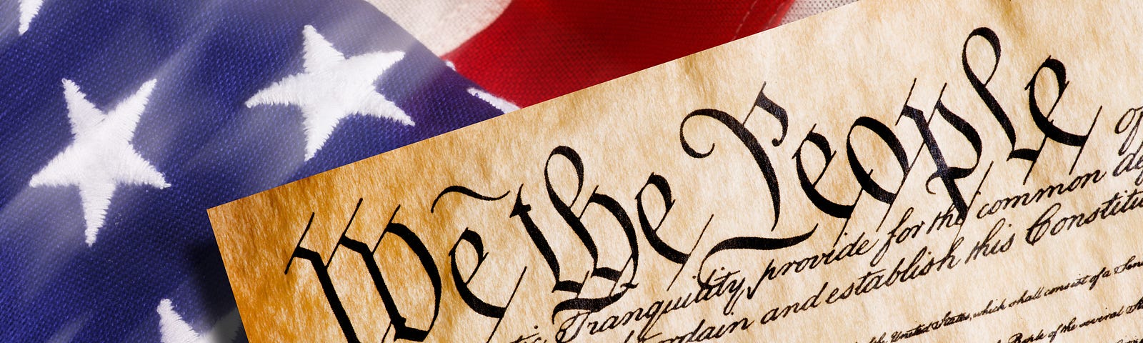 Close-up of an image of the Constitution showing the “We the People” heading. It is laying atop a folding American flag.