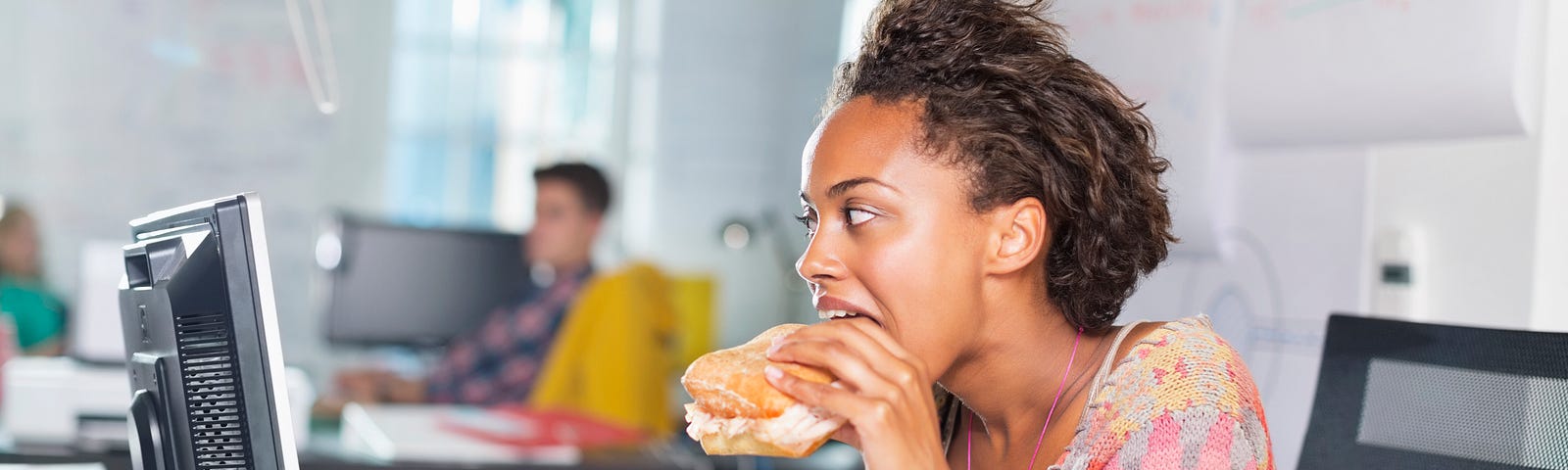 A businesswoman eating a burger at her desk as she scrolls on her laptop.