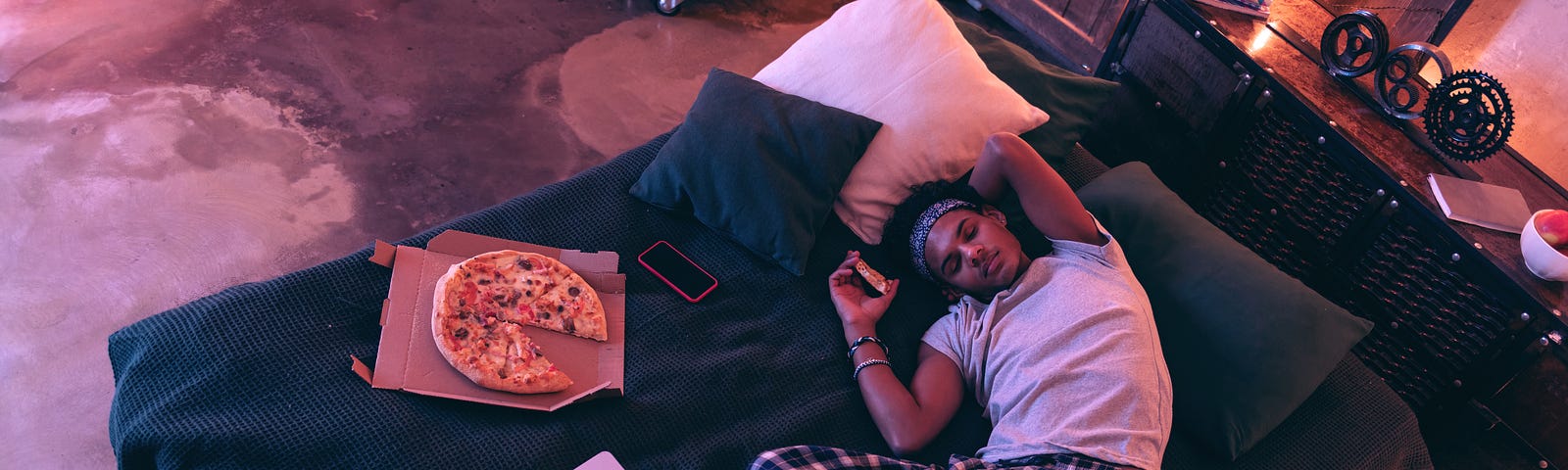 A young guy in checkered pajama trousers falling asleep with a slice of pizza in his hand in bed next to a pizza box and open laptop. He has the itis.