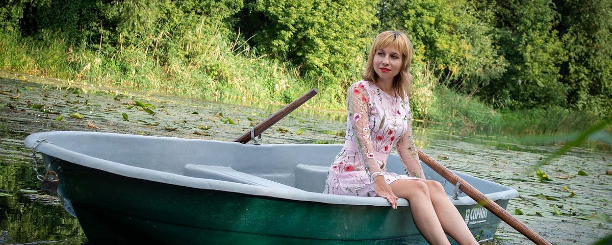 An attractive woman sitting in a rowing boat on a small, quiet river. Her legs are dangling over the side of the boat and her bare feet are trailing in the water.