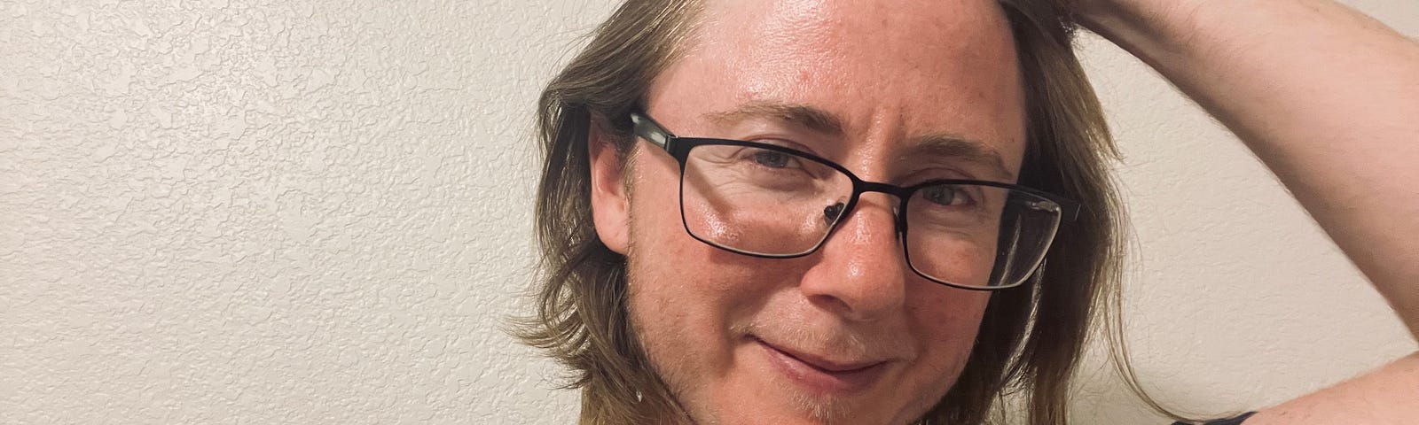 Author is wearing a t-shirt and black framed glasses. He is holding his hair back and has the beginnings of a beard. He’s smiling just a little.