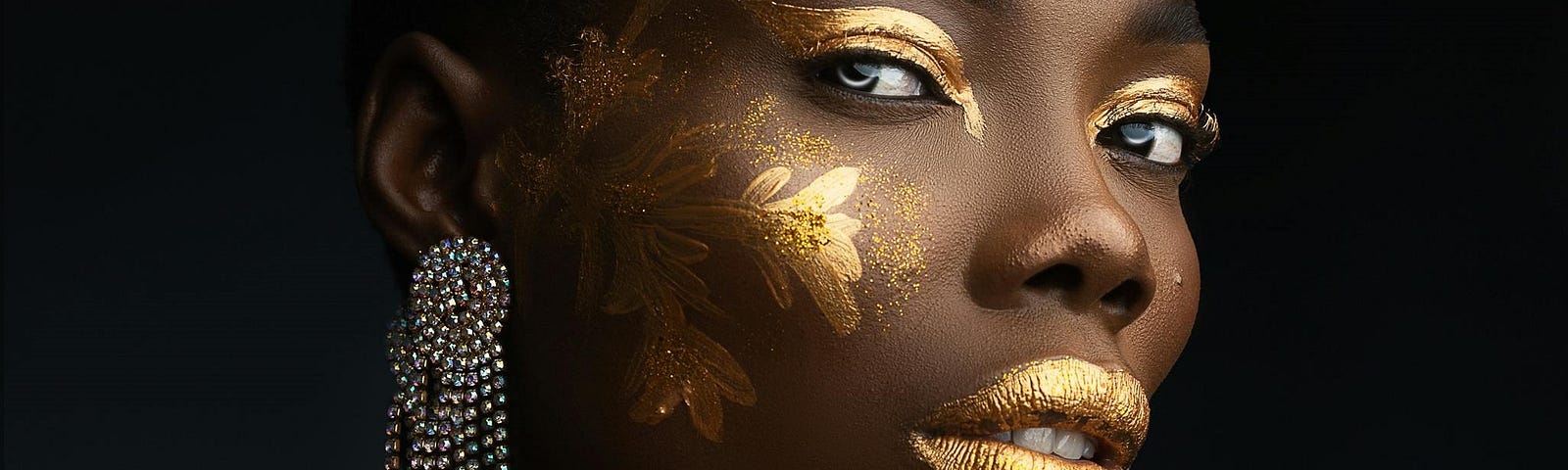 A woman of African descent stares at the camera in a close-up shot, there are flowers painted on the right-side of her face in gold makeup, and her eyeshadow and lips are colored gold. She’s wearing a neck bangle and long, silver earrings.