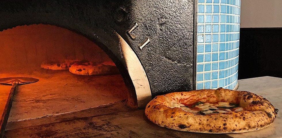 A baked pizza sitting on the hearth of a pizza oven that currently has two other pizzas in it.