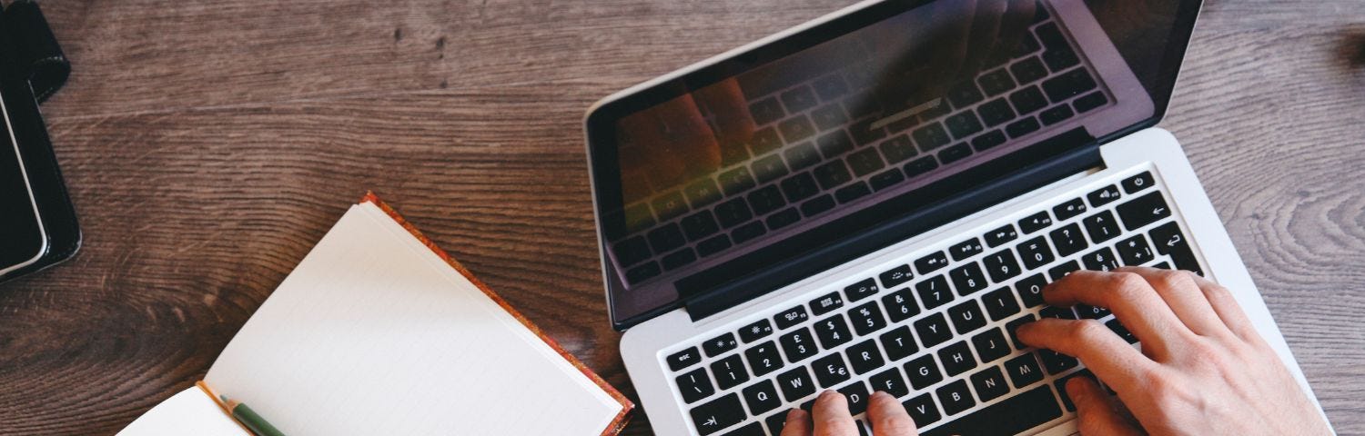 Image of hands typing on a laptop keyboard with a notebook to the person’s left.