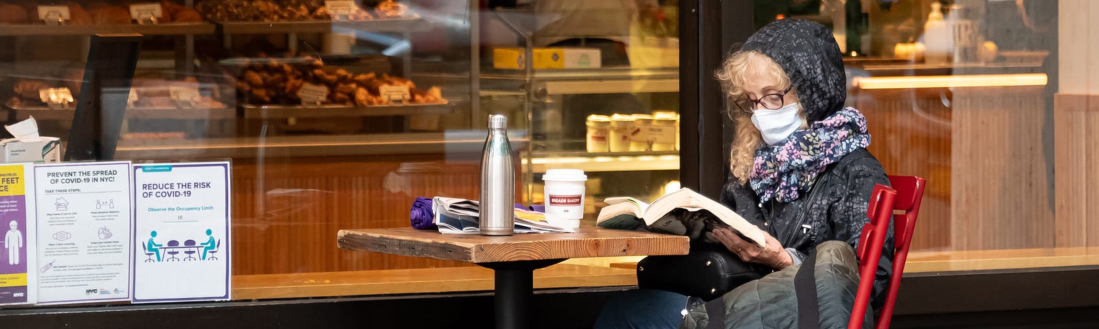 Older woman wearing face mask reading a book outside a cafe.