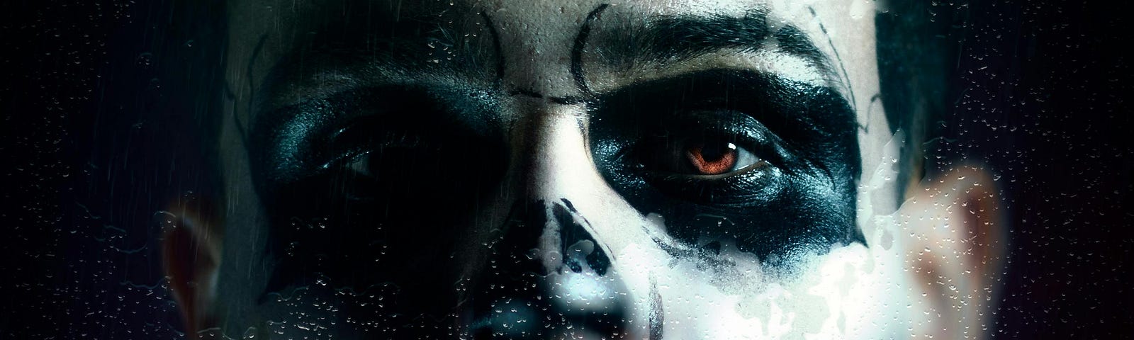 A close-up shot of a man with ghostly black and white face paint, he’s staring through a water-soaked window. The image is focused on his left brown eye.