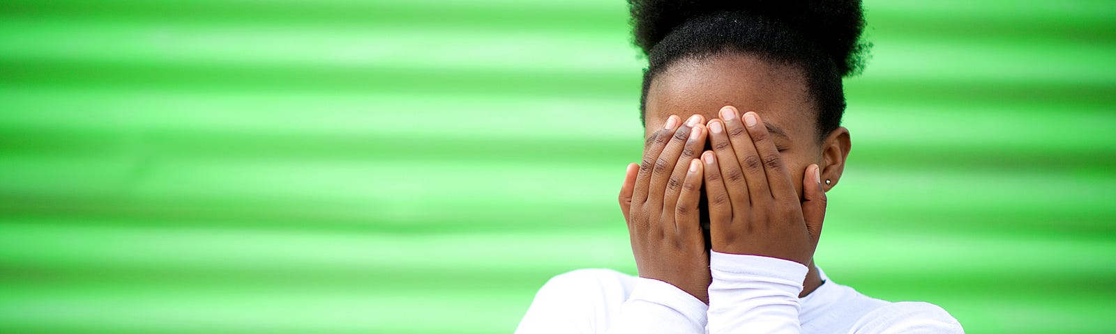 A girl covers her face with her hands against a bright green background.