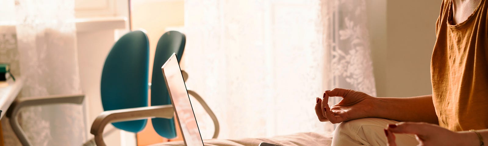 A woman in white jeans and a yellow sweater sitting on the bed in a yoga pose in front of a laptop and a cup of coffee.