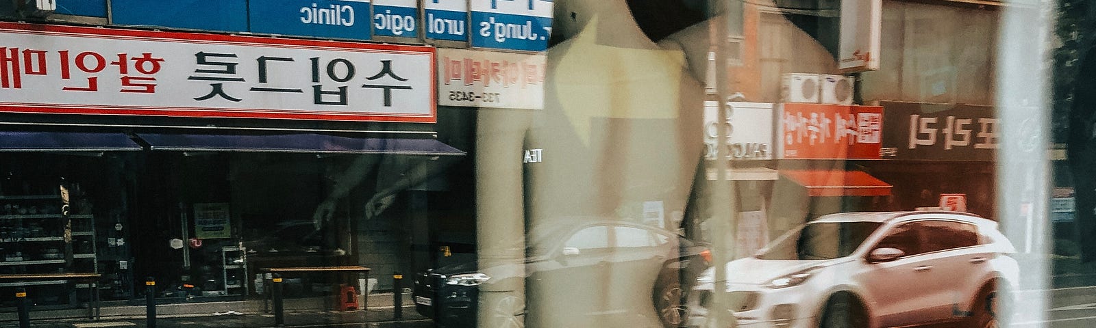 A window reflecting a car driving down the street and Korean shop signs are two naked white mannequins facing away from the viewer.