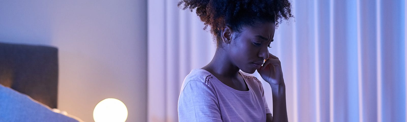 A troubled black woman sits at the edge of her bed.