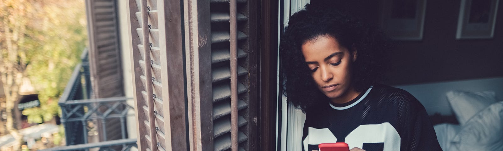 A photo of a black woman on her phone looking out of her apartment window/door.