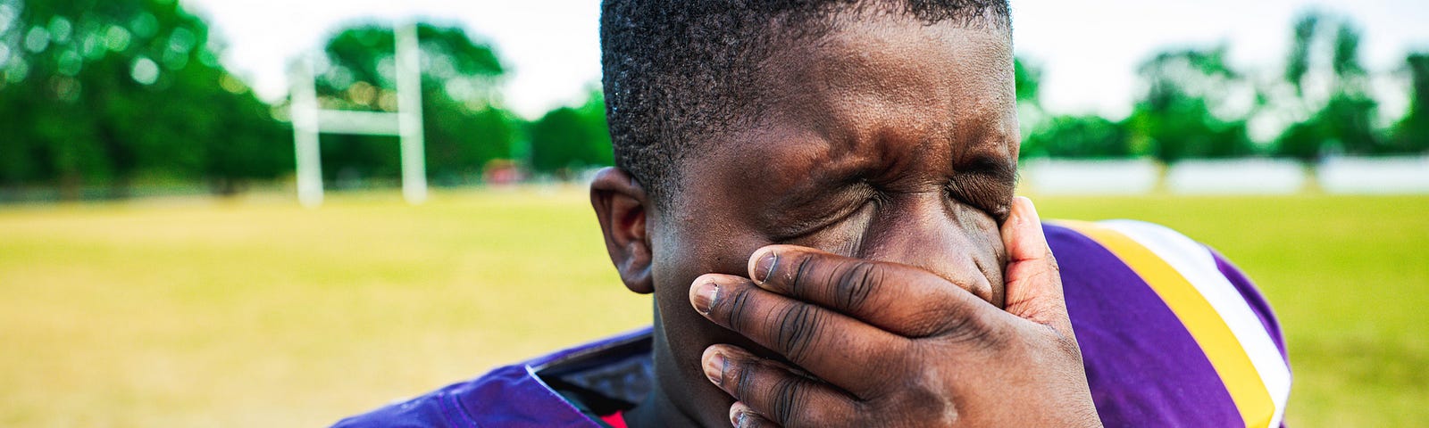 A close-up of a young football player rubbing his face with his hand in exhaustion.