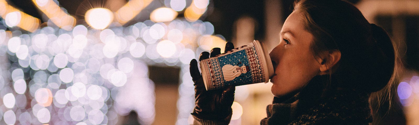 Woman drinking a coffee in a Christmas design cup with Christmas lights in background.