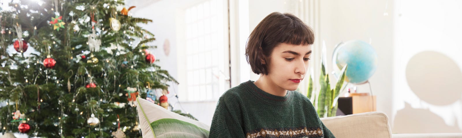 A person with short bobbed hair sitting on a couch in front of a Christmas tree, working on a laptop.