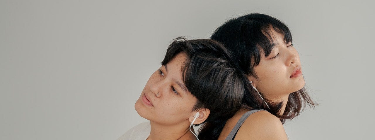 Picture of two women sitting on a bed, back to back, one of them with auriculars, the other playing guitar