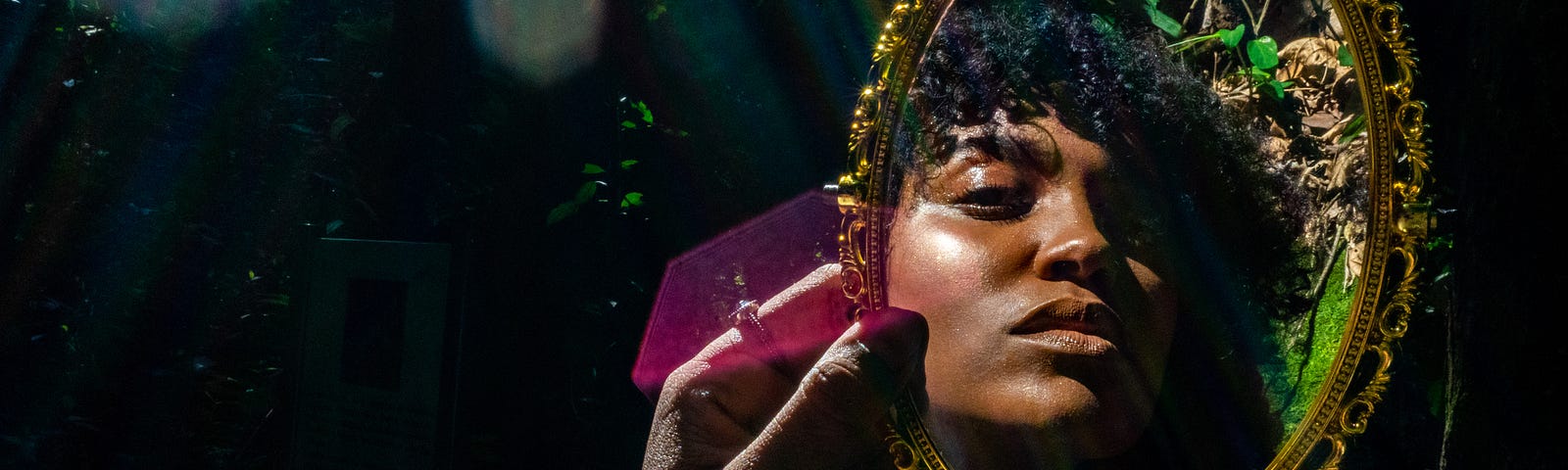 Young Black woman holding up and looking into a gilded mirror in the forest.