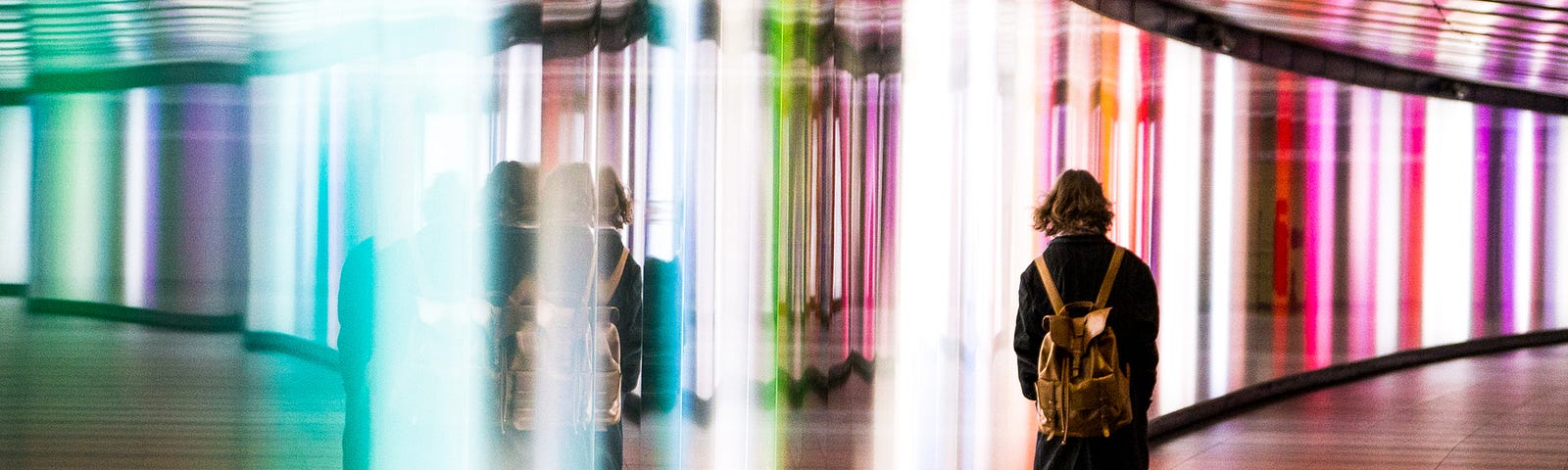 A photo of a woman with a backpack on walking through a bright rainbow-lit curved hallway.